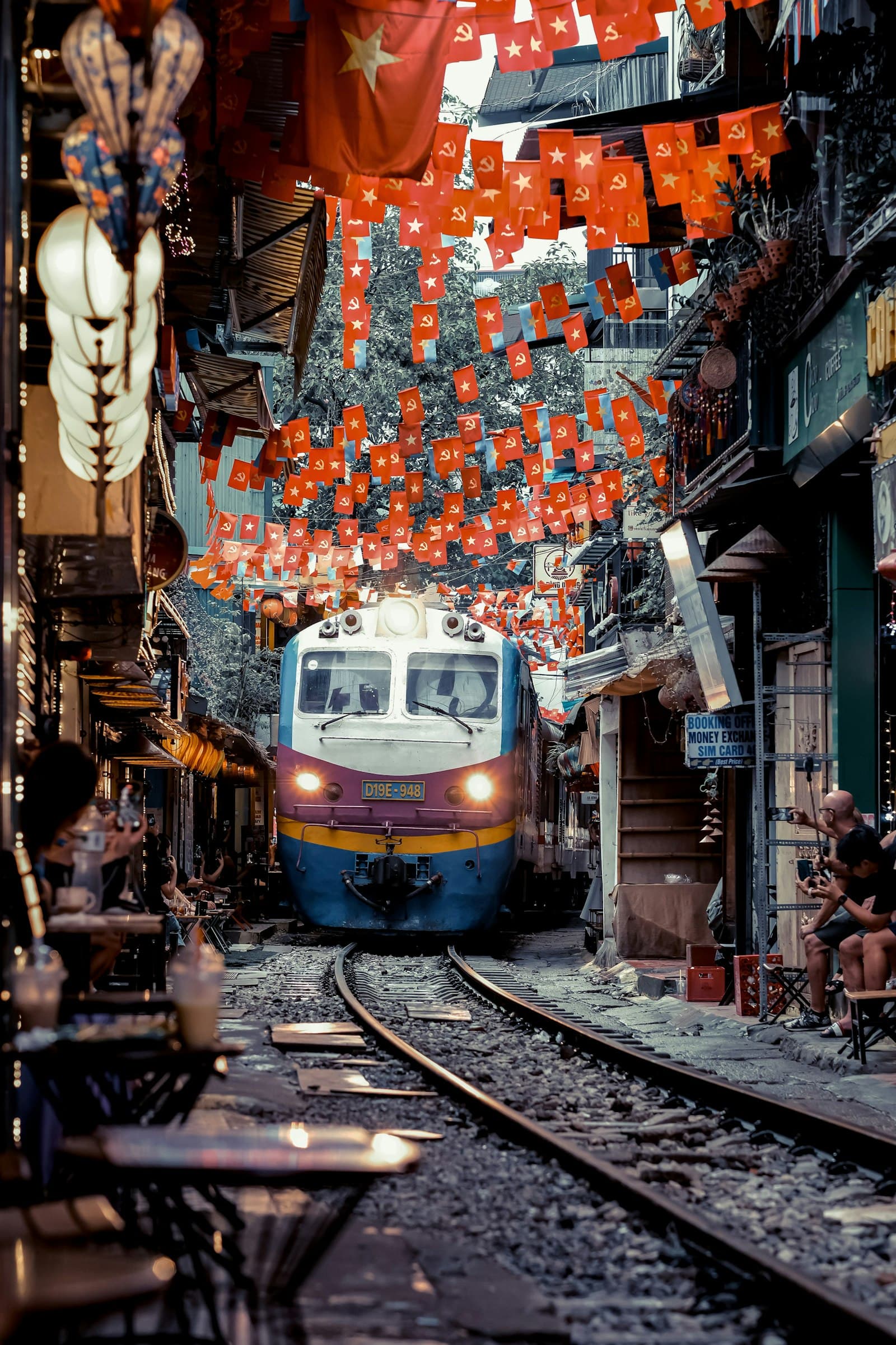 The Reunification Express train easing through Hanoi Train Street with red flags overhead